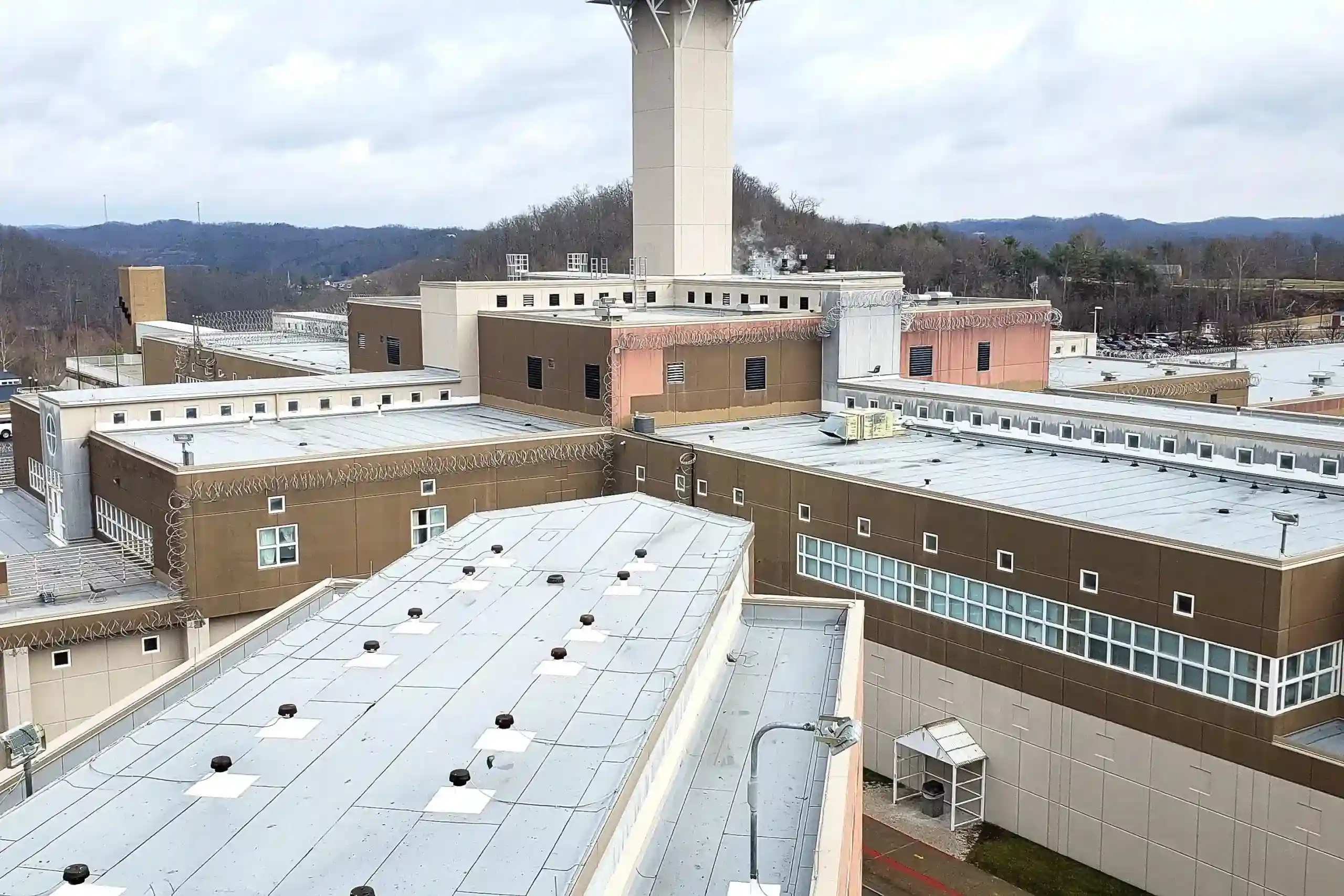 Aerial view of a correctional facility with razor wire-topped rooftops and a central guard tower, surrounded by wooded hills.