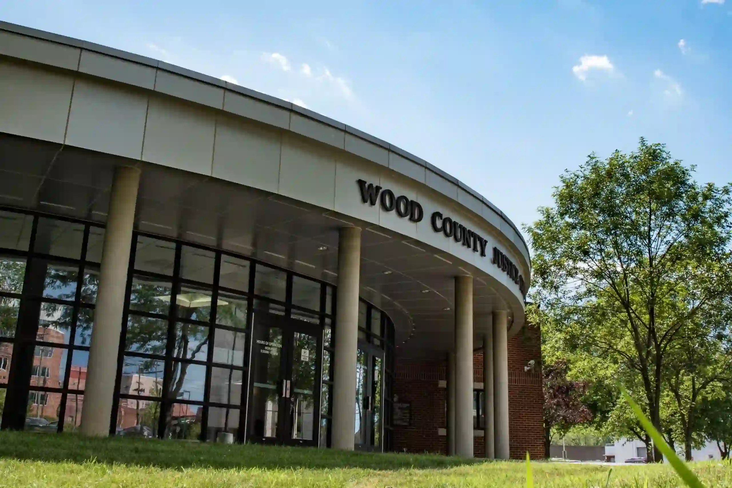 Low-angle view of the Wood County Justice Center entrance, showing a curved metal-clad overhang, brick and glass facade, and columned walkway surrounded by trees on a sunny day.