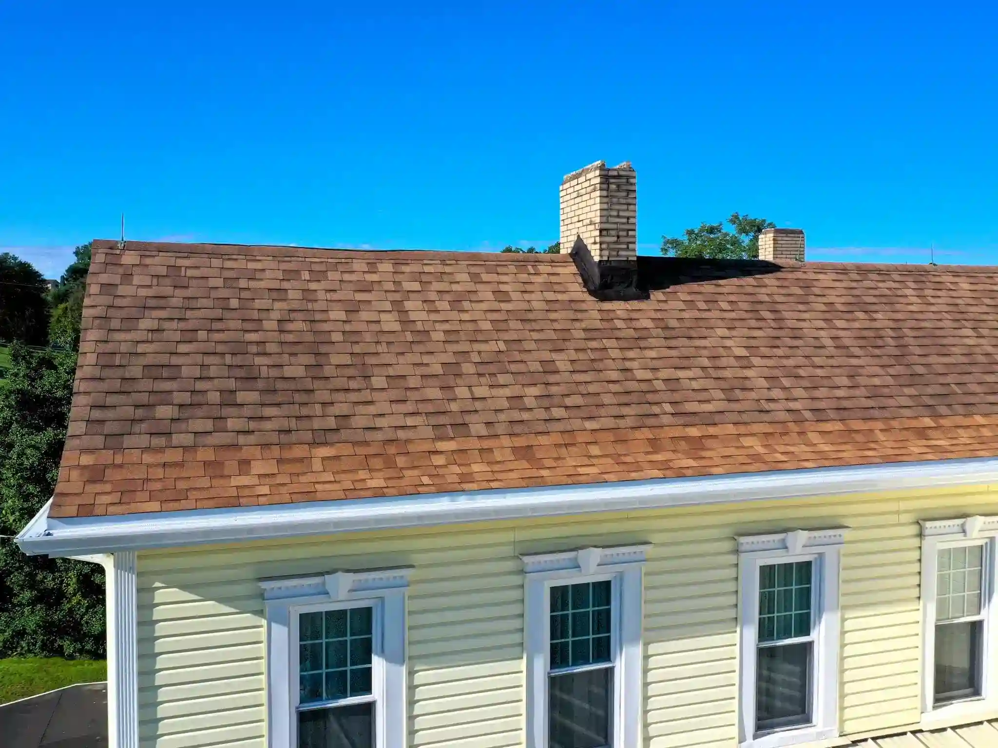 Aerial view of a brown asphalt shingle roof with brick chimneys and white gutters on a house with pale yellow siding.