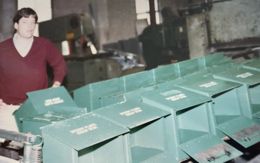 Vintage photograph of a worker standing beside a row of green metal "Speedy Plate Drop Box" bins on a factory floor, with industrial equipment visible in the background.