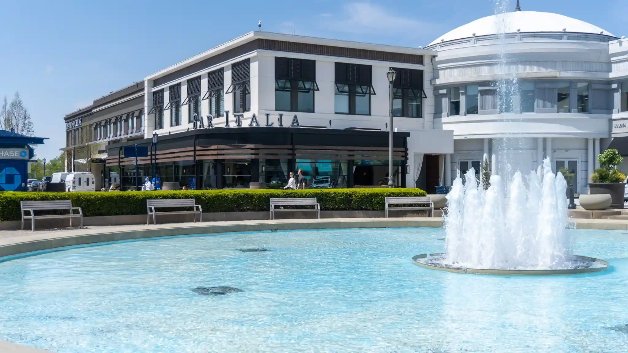 Outdoor plaza with a large circular fountain spraying water in the foreground, benches and hedges lining the pool, and a row of upscale shops and restaurants, including Bar Italia, in a white and dark-trimmed building with a domed rotunda under a clear blue sky.