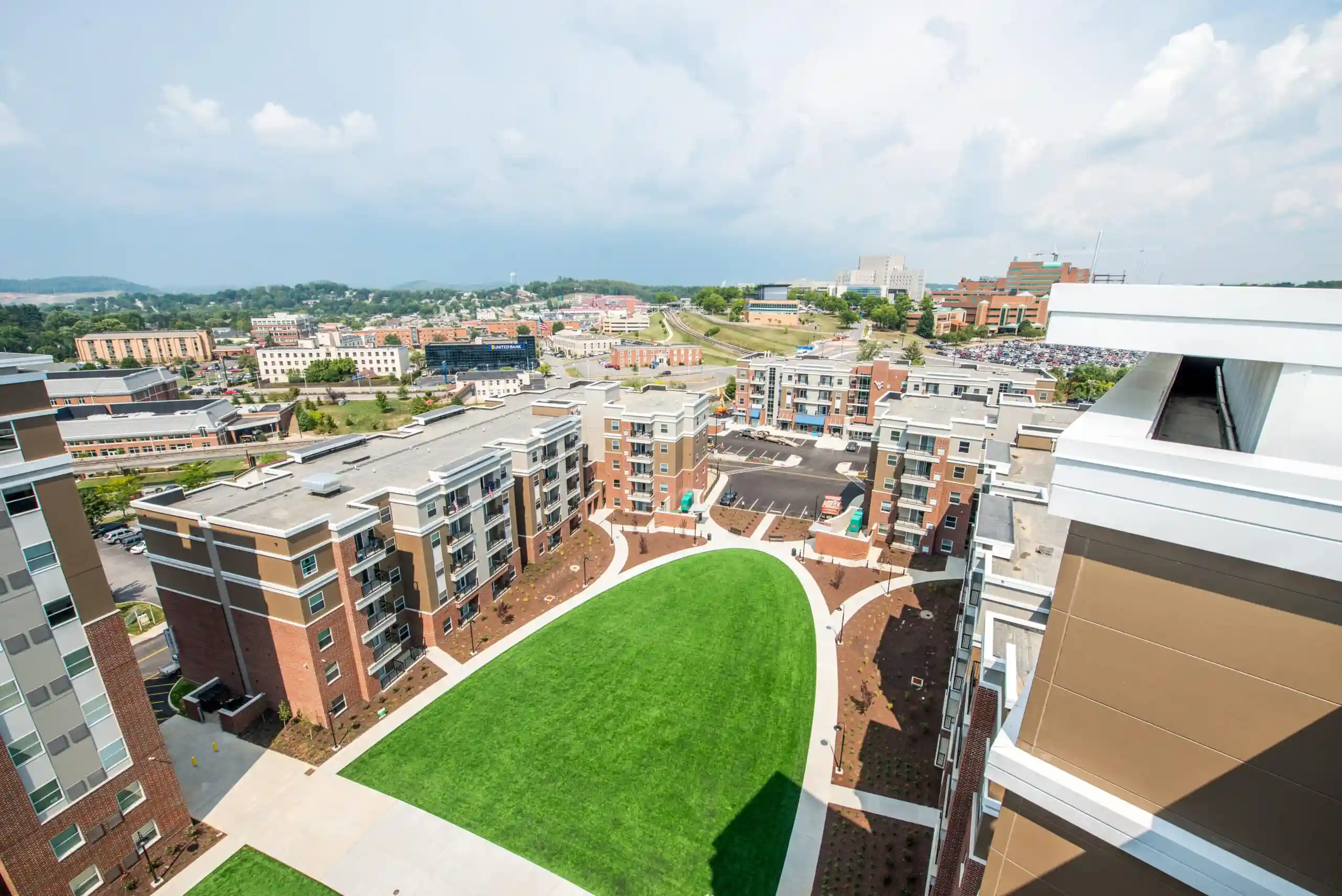Aerial view of a modern apartment complex with brick and beige mid-rise buildings arranged around a curved central lawn and courtyard, with a hospital campus, city buildings, and wooded hills visible in the background.