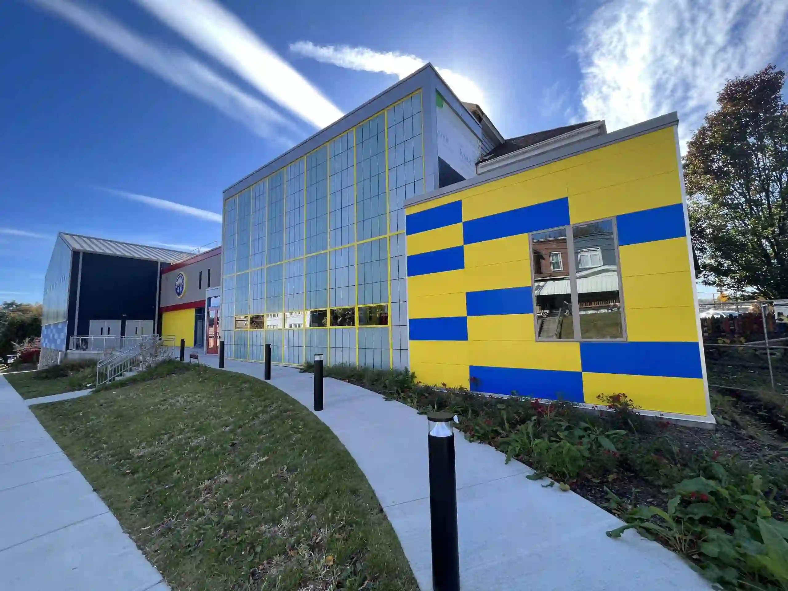 Exterior of a modern building with a bold yellow-and-blue checkered facade, a glass curtain-wall section with yellow mullions, and a connected navy metal-roofed gymnasium, set on a landscaped hillside with a curved sidewalk under a bright blue sky.