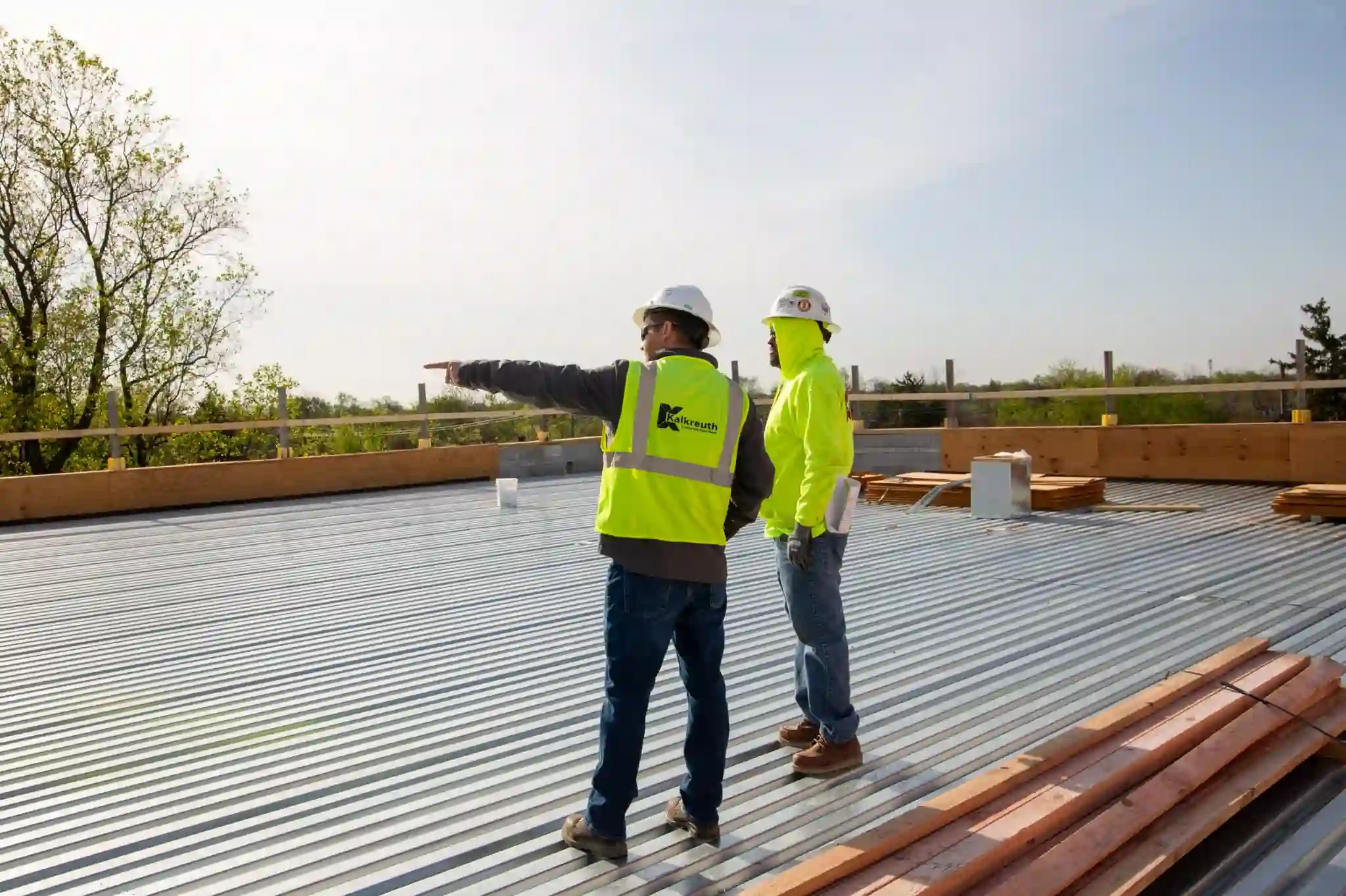 Two construction workers in hard hats and high-visibility gear stand on a metal roof deck under construction