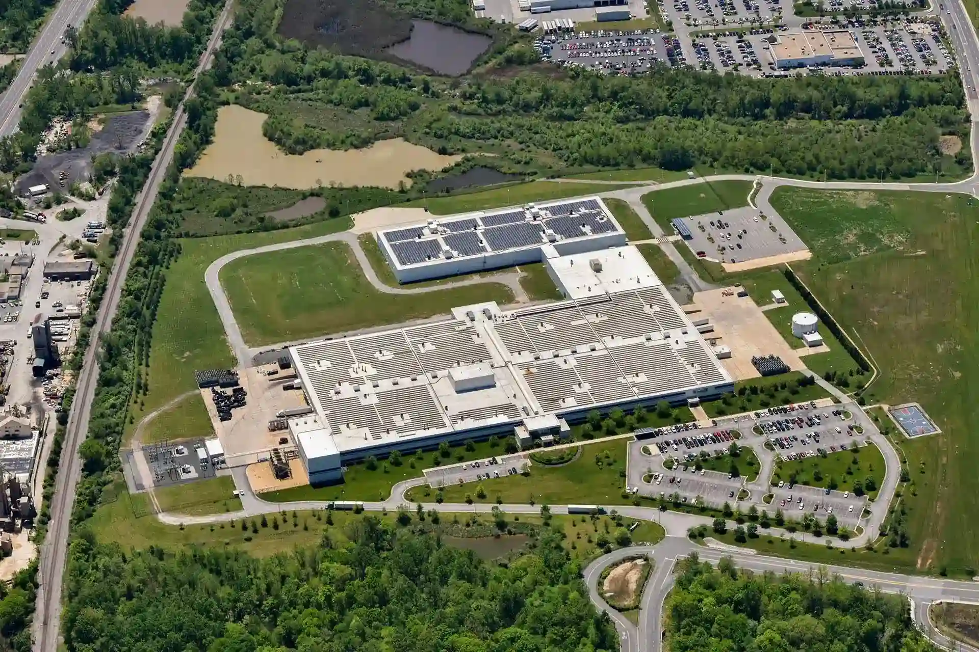 Aerial view of a large industrial or manufacturing facility with a white flat roof, including a smaller adjacent building topped with solar panels.