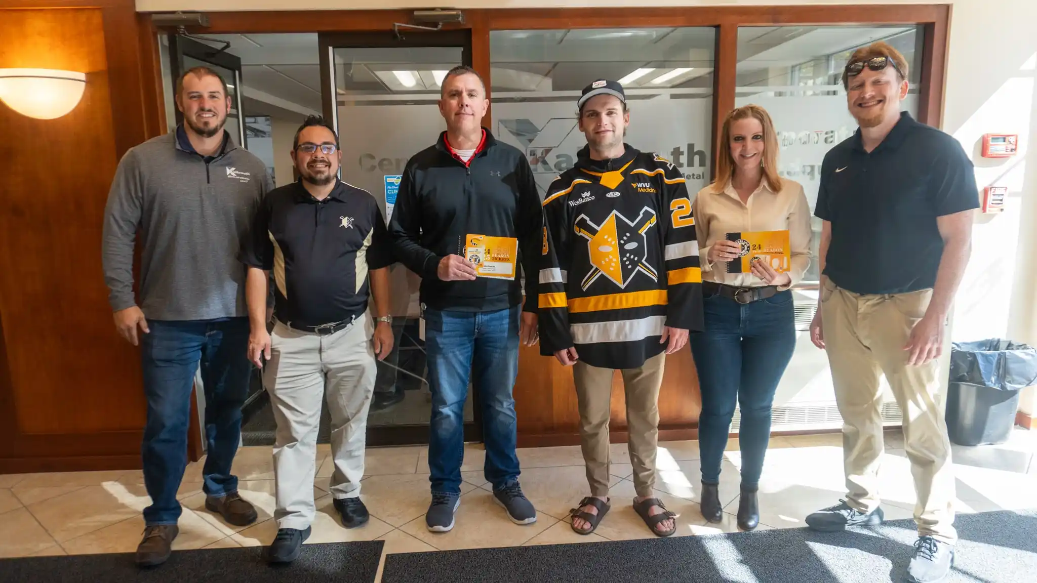 Six employees pose in a corporate lobby, with two holding season ticket vouchers and one wearing a black and gold hockey jersey.