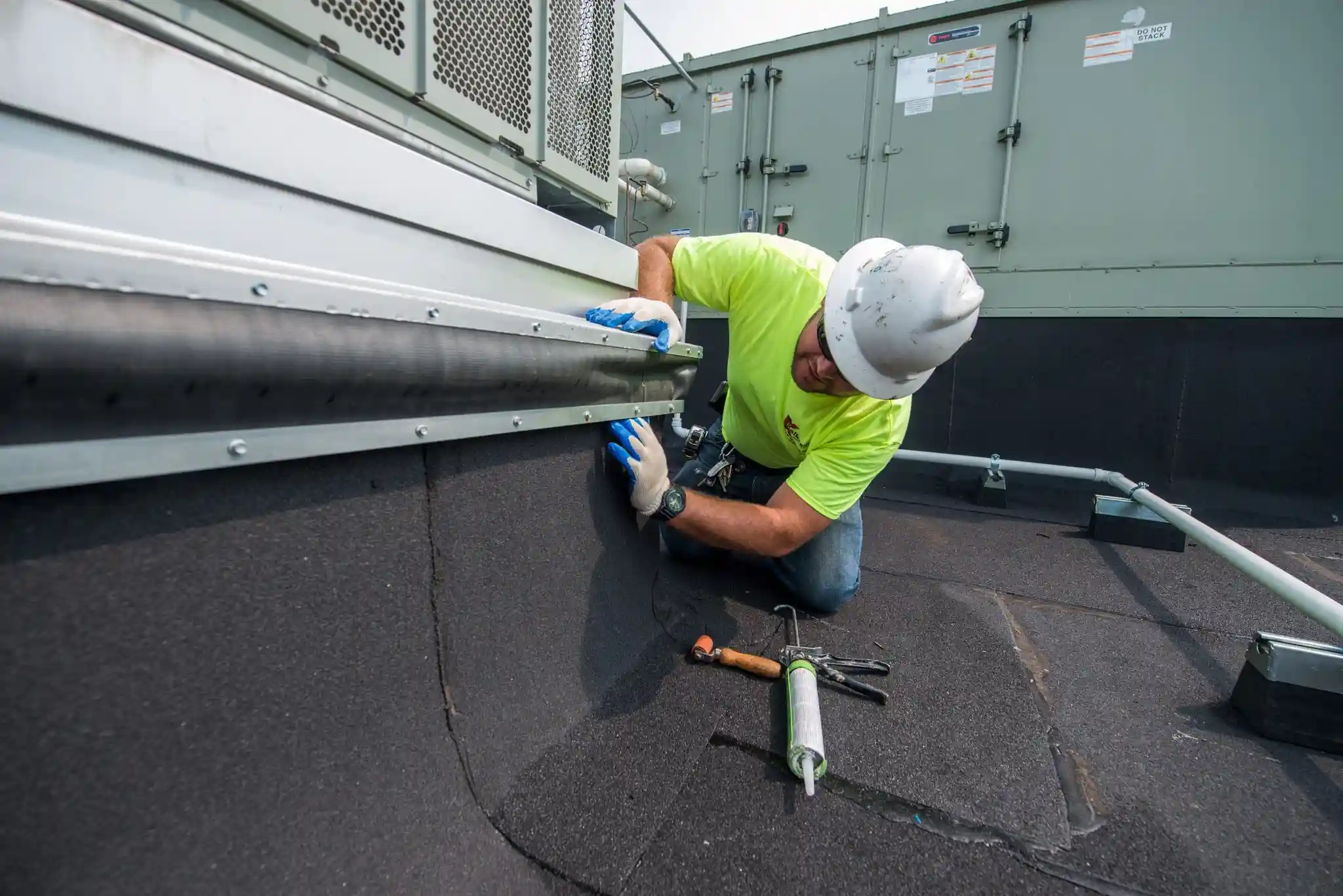 A technician in a hard hat and safety gloves kneels on a flat commercial roof, inspecting and sealing.